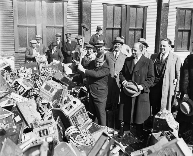Fiorello LaGuardia smashes slot machines with a sledgehammer while a crowd watches on.