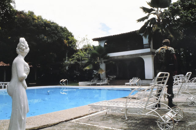 A statue of a woman with a swimming pool behind it. A Spanish colonial house behind that, as a soldier walks along the poolside.