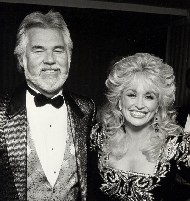 Black and white photo of Dolly Parton and Kenny Rogers smiling, both in sparklingl clothing.
