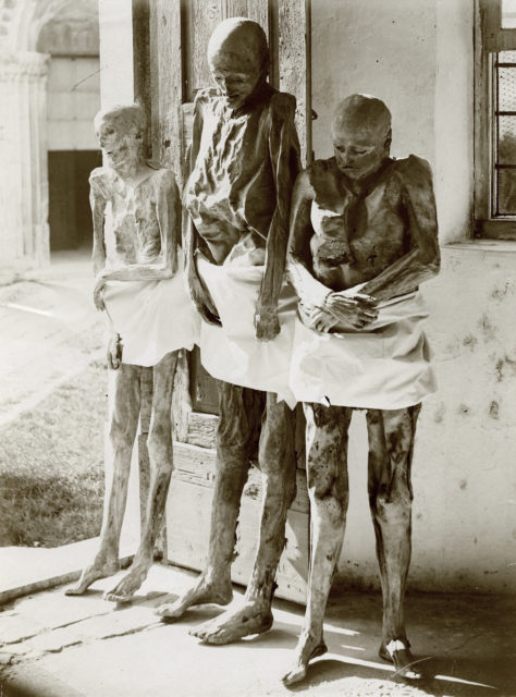 Three naked mummies standing tied to a building post.