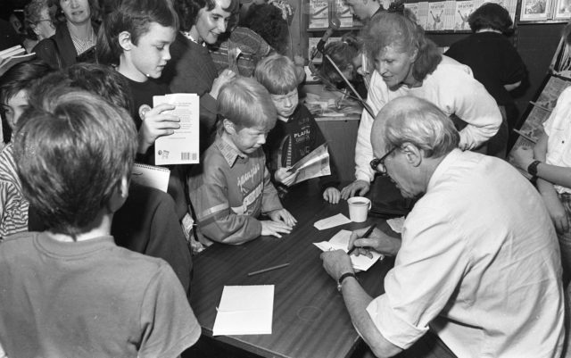 Roald Dahl sitting at a table signing books brought to him by several children surrounding the table.