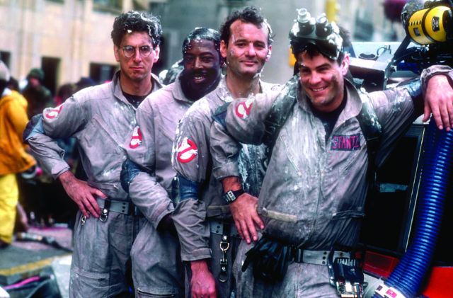 Harold Ramis, Ernie Hudson, Bill Murray, and Dan Aykroyd posing in a line against a vehicle in costume from 'Ghostbusters'