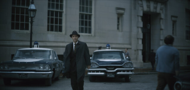 Alessandro Nivola in a jacket and hat walking away from two police cars.