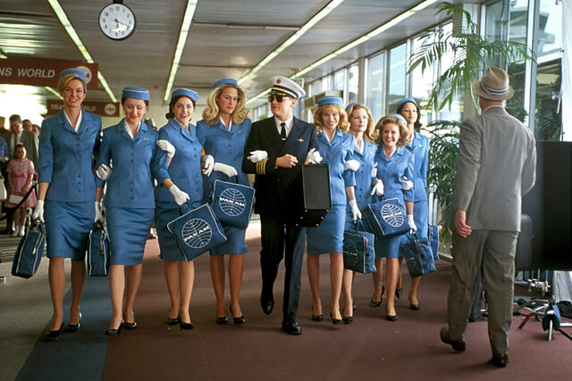Leonardo DiCaprio in a pilot's uniform walking arm in arm with a group of flight attendants in blue.