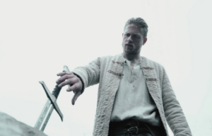 Charlie Hunnam in a white jacket reaching down towards a sword.