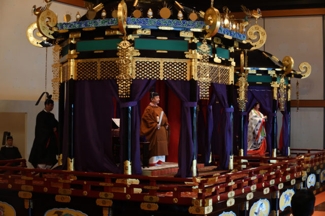 Emperor Naruhito stands in the chrysanthemum throne during his enthronement ceremony, his wife also on the throne but further away.