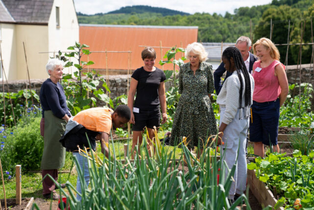Camilla, Queen Consort, and King Charles stand in a garden with a group of people.
