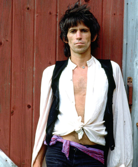 Keith Richards leans up against a red wall wearing a white button down shirt with a vest