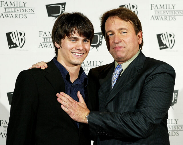 John and Jason Ritter smiling for a photo, John has his hand on his son's shoulder and chest.