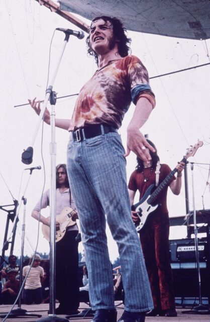 Joe Cocker in striped pants and a tie dye shirt singing into a microphone. 