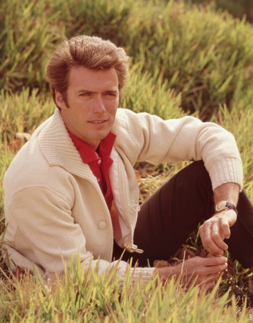Portrait of American actor and director Clint Eastwood sitting in a field, with his leg bent and his elbow resting on his knee. Eastwood is wearing a white cardigan over a red shirt.
