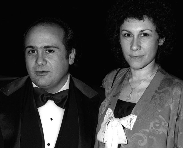 Young Danny DeVito in a suit and Rhea Perlman in shirt with a white bow on the front.