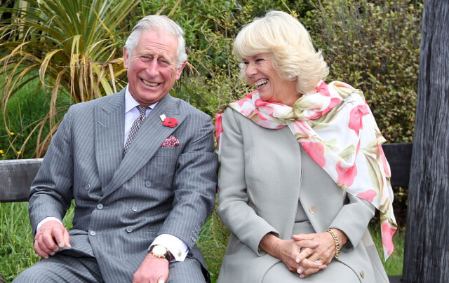 Camilla and Charles in matching grey outfits, laugh together while sitting on a bench.