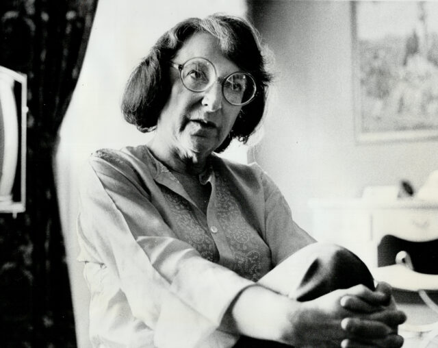 A photo of Pauline Kael sitting with her hands wrapped around her crossed legs.