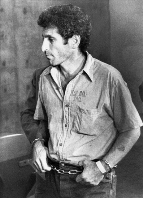 Angelo Buono in handcuffs walking through a hallway.