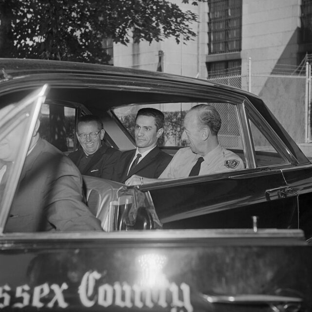 George Nassar in a suit in the back of a car sitting between two police officers. 