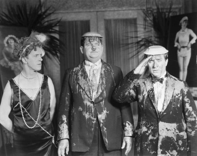 Stan Laurel, wearing a dress, looks at Oliver Hardy and another actor covered with pie in a movie still. 
