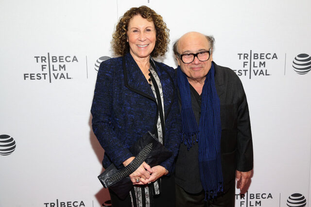 Rhea Perlman in a blue jacket and Danny DeVito in a black shirt with blue scarf posing on the red carpet together.