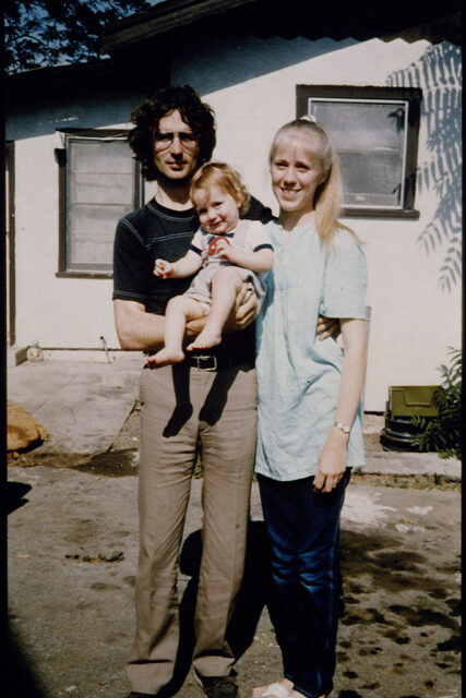 David Koresh holding son Cyrus with his arm around his wife Rachel as they all smile for a photo in front of a white house.