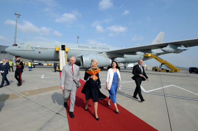 Charles and Camilla walk down a red carpet away from a plane with an escort. 