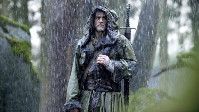 Charlie Hunnam carrying a sword with a cloak over his head standing in a rainy forest. 