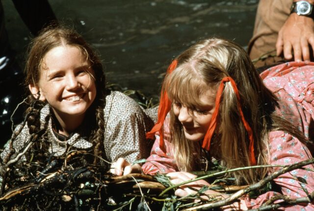 Young Melissa Gilbert and Alison Arngrim in dresses smiling at the camera while they lie on their stomaches.