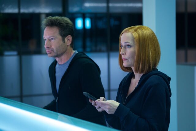 David Duchovny and Gillian Anderson, who is texting, stand in an office wearing black sweaters.