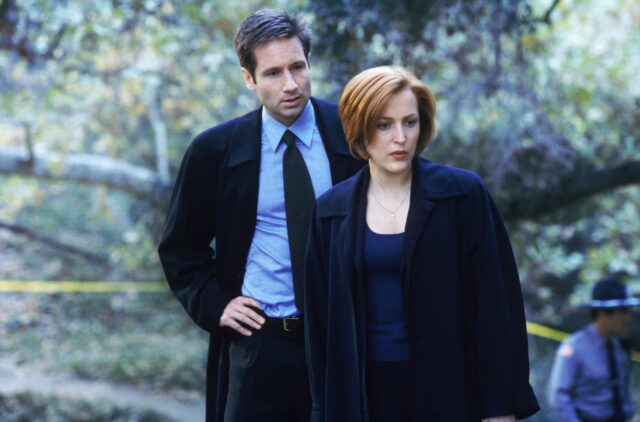 Gillian Anderson and David Duchovny in black suits and jackets surrounded by forest. 
