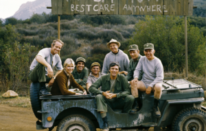 Cast of M*A*S*H posing together on a jeep in front of their camp sign.