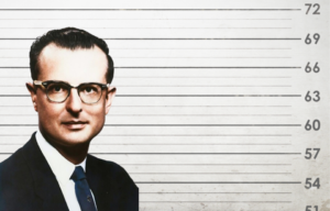 John List in a black suit and glasses in front of a mug shot wall.