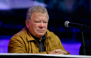 William Shatner sitting with his hands folded in front of a microphone, looking sad.