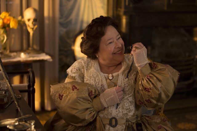 Kathy Bates as Delphine LaLaurie in a beige dress with puffed sleeves lifting her hand up to her face.