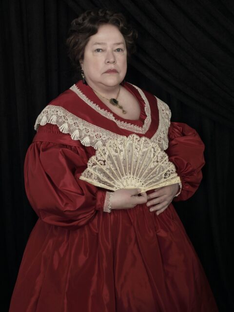 Kathy Bates as Madame LaLaurie in a red dress with puffed sleeves, a white lace collar, and a fan. 