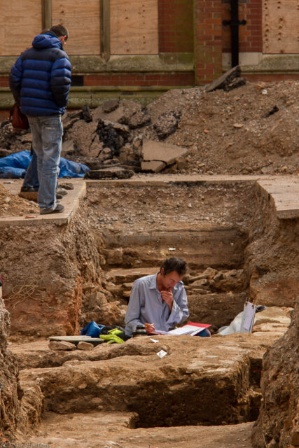 Archaeologist writing notes in a trench while another stands above him