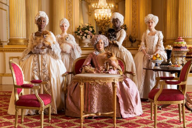 Group of women in regal gown and wearing powdered wigs stand around another woman who is seated.