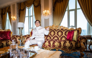 Jackie Chan in a cream tracksuit sitting on an elegant gold couch pointing to the side.