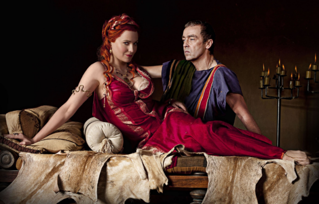 Lucy Lawless in a red dress, and John Hannah in a toga, sit beside each other on a Roman bed
