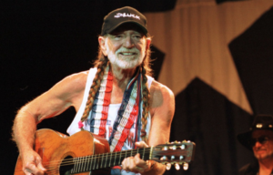 Willie Nelson playing guitar while wearing a white tank top, black baseball cap, and two braids in his hair.
