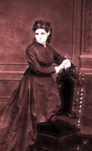 Madame LaLaurie kneeling on a chair with her arms on the back, wearing a red dress.