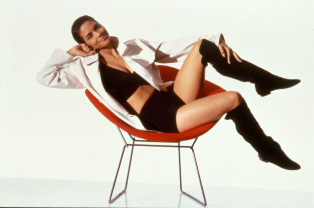 Young Halle Berry sitting sideways on a chair in a black tank top, shorts, and a long sleeved white shirt.