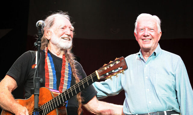 Willie Nelson and Jimmy Carter smiling with their arms around each other on stage.
