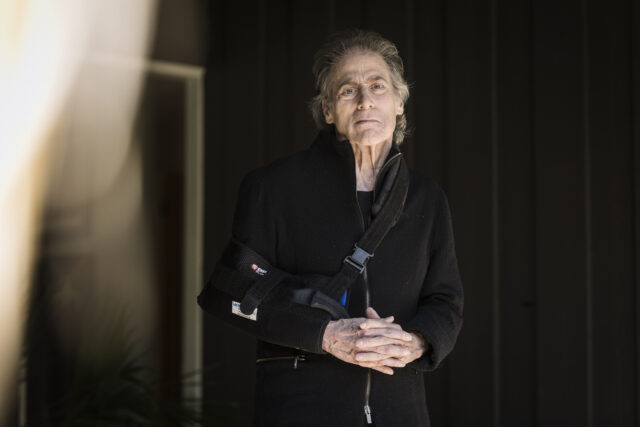Richard Lewis standing with his arm in a sling, hands clasped in front of his stomach.