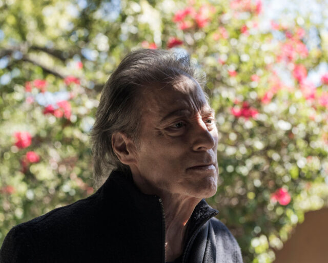 Headshot of Richard Lewis with shadows on his face, standing in a garden.