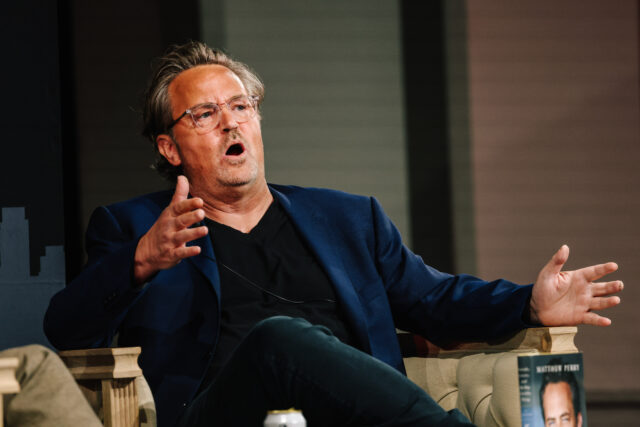 Matthew Perry speaking with his hands out in front of him while seated.