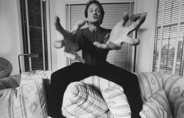 Robin Williams standing on a couch, hands reaching toward the camera.