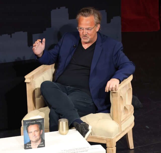 View of Matthew Perry sitting on a chair, a table in front of him holding his memoir on display.