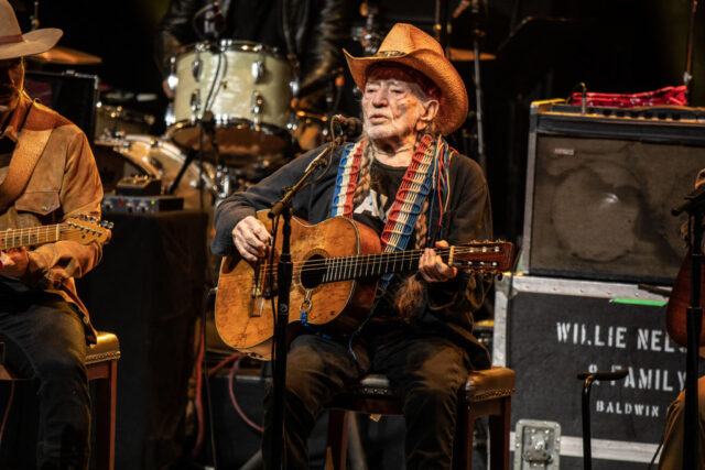 Willie Nelson sitting on stage playing a guitar wearing a black shirt and a cowboy hat.