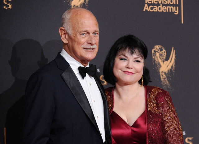 Gerald McRaney in a black suit with a bow tie, and Delta Burke in a red and gold dress, pose for a photo together on the red carpet. 