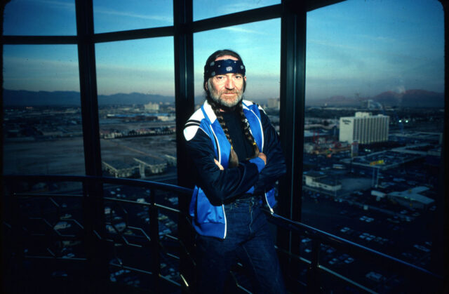 Willie Nelson in a blue vest with his arms crossed, and his hair in two braids. 