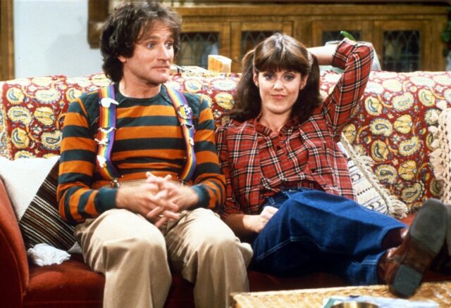 Robin Williams and Pam Dawber sitting on a couch while filming for the sitcom "Mork & Mindy."